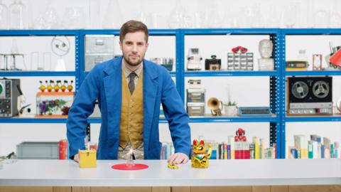 GCSE physics image: a presenter in a blue lab coat and waistcoat stands at a table in a workshop with various scientific devices.