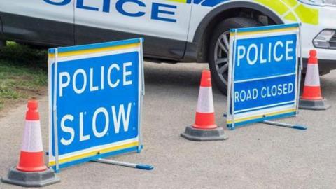 Two blue police signs, one saying "police slow" and the other saying "police road closed", stand on the road with one traffic cone between them and one on either side of them. 