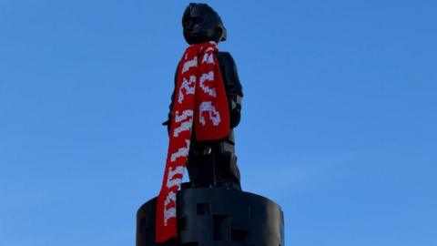 A shiny black statue of a small child, who looks as if they would be about 6 or 7 years old. A Barnsley FC scarf is draped around his neck.