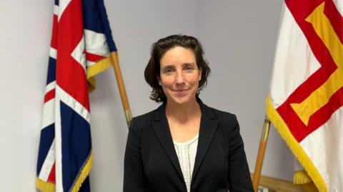 This is a photo of Deputy Lindsay de Sausmarez. She is a woman with medium length dark hair. She is smiling at the camera, wearing a white blouse with a black jacket. She is standing inside and there are two flags behind her.