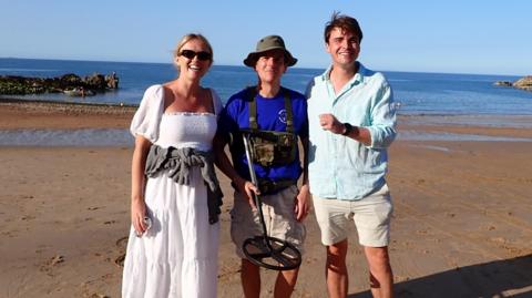 Alexandra Lee with Steven Andrews and Ollie Lee on a beach. Alexandra is wearing a white dress while Steven is wearing a blue top and is holding his metal detector. Ollie is wearing a blue shirt. They are stood on the sand and the tide is out. 