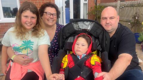 Rob Channon (right) kneels next to his son Gethin, who sits in a wheelchair. Rob is bald and has brown eyes and wears a black polo shirt. Sian Channon kneels on the left of the wheelchair, she smiles at the camera and has short brown hair and has black rimmed glasses. Their daughter sits on Sian's knee on the far left. She has mousy brown curly hair and smiles at the camera. The back of their house is seen behind them. 