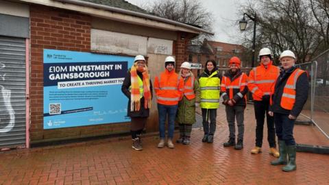 People in hi-vis jackets and hard hats standing next to the former toilet building. There is a blue poster on the wall of the building providing details about investment in the town, along with a grey metal roller door.