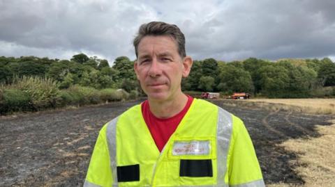 A man with brown hair standing in front of a burnt field in a yellow hi vis jacket 
