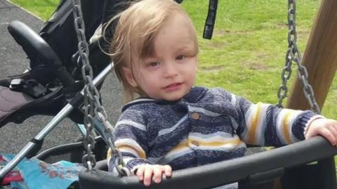 Bronson Battersby, wearing a blue and yellow cardigan, on a swing with a pushchair in the background