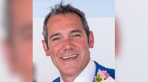 Jeremy Clark smiles at the camera. he is wearing a suit and white shirt with a floral buttonhole on his jacket.