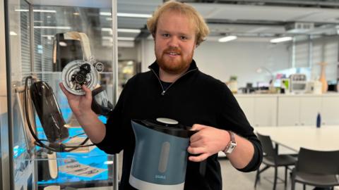 A man in a black top with the sleeves pulled up holding his repairable kettle in the design lab of De Montfort University