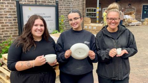 Three women are wearing black, holding white crockery and stand in front of a wooden pallets, cardboard boxes, plants, a brick bottle kiln and a sign reading 'Welcome to Dudson Museum'. The woman on the left has a tattooed arm, long brown hair and is holding a cup. The woman in the centre wears glasses, earings, a black jumper with a white logo on the left chest and is holding a white plate. The woman on the right has blonde hair tied in a bob, glasses, a black waterproof jacket and is holding a small white milk jug.