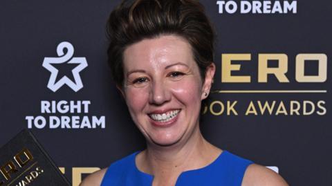 Claire Lynch smiling in blue dress in front of Nero Book Awards branding