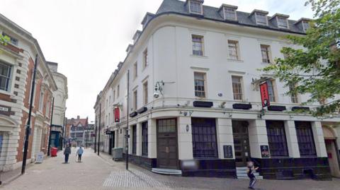 A view of a white multi-storey building on the corner of a street, next to a pedestrianised walkway.