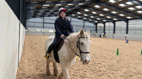 A woman wearing dark-coloured clothing is pictured riding a white horse in a large indoor riding arena.