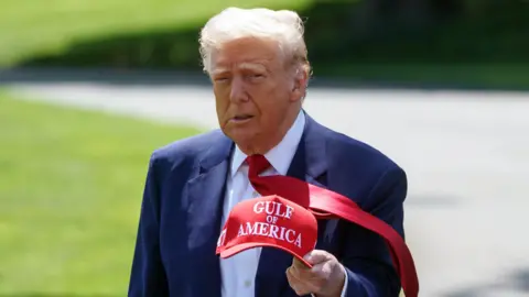 Donald Trump wearing a blue suit holding a red hat saying 'Gulf of America'