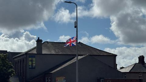 A picture of the lamppost with the union jack flag attached. The flag is billowing in the wind about half way up the lamppost. It has been hung upside down. Above it, a CCTV camera has been attached to the lamppost. A church stands behind the lamppost and has a poster displaying a cross on its wall.
