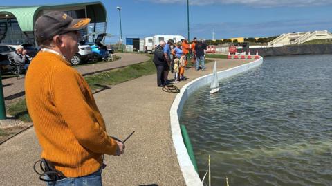 A member of Redcar Model Boat Club is sailing a mini vessel in the refilled section of the lake. He is wearing an orange jumper, jeans and a black cap. A white mini boat is sailing in the water. A group of people, including two children are looking on. There are parked cars on the grassland next to the lake. 