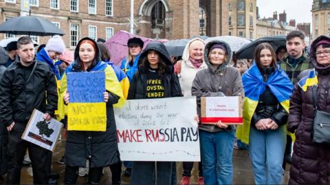 A group of around a dozen people stand together holding up hand-drawn protest signs, one reading 'make Russia pay' in red and black. They are all wearing raincoats and some are draped in Ukrainian flags. They are standing in front of City Hall in Bristol, a large brown brick building, on a grey, rainy day.