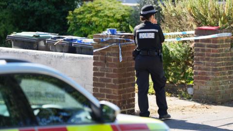 A police officer stands at the walled entrance to a footpath towards a house. The path is cordoned off with police tape. 