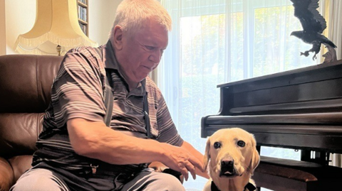 Graham Grange is sat on a brown leather chair and is tending to Lylah, a gold-coloured dog, who is sat on a rug on the floor, in front of a picture 