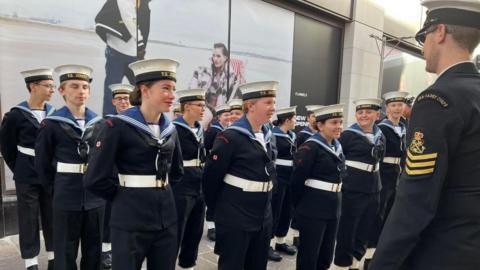 A group of young sea cadets stand at attention in uniform in front of a commanding officer