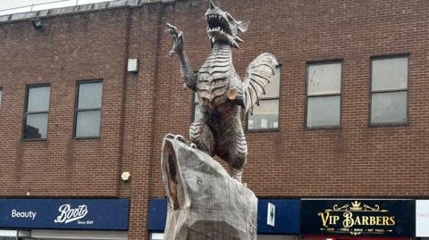 The picture shows the wooden sculpture in Taunton.  The dragon's arm and tongue are missing. There are shops in the background.