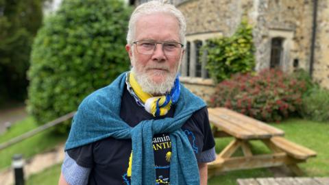 Victor Ient with glasses and a grey beard standing in a garden, wearing a scarf and sweater over the shoulders, with a picnic table and stone building in the background.
