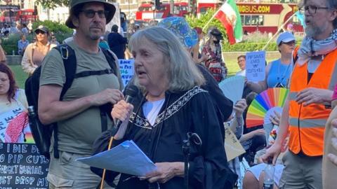Joy Dove speaking at the rally on Parliament Square. She has shoulder-length grey hair and is holding a microphone in one hand and her speech in the other. A sign language interpreter stands to her left. She is surrounded by protestors holding placards.