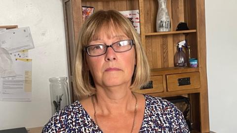 Wendy Randall pictured alone wearing glasses and a long sleeved top. She is also wearing a silver necklace and is standing in a home office environment.