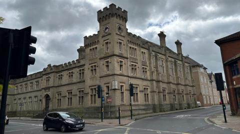 A view of the exterior of County Hall in Ipswich. The building is made of large grey bricks and has a similar look to a castle. People can be seen walking past.