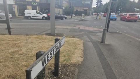 A busy road with traffic from both directions, coming out from a side street and cars parked up. The road sign identifies the street name as Westgate. Shops in the background suggest it's a busy trade route.