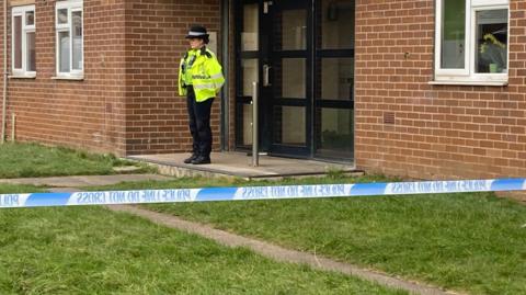 A female police officer wearing a yellow jacket over her black uniform and hat stands in front of the entrance to a block of flats