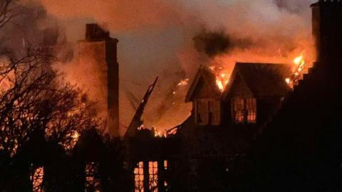 Flames engulf a large building at night, with thick smoke billowing into the sky as fire spreads through the roof and windows.