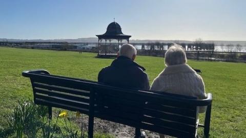 A wide grassy park on a sunny day, with two people sitting on a black bench facing away from the camera. In the distance stands an ornate bandstand and a large body of water. Daffodils bloom near the bench, and the sky is clear and bright.