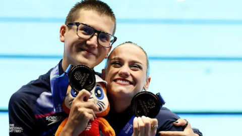 Ranjuo Tomblin and Izzy Thorpe stand side by side with their arms around each other in front of a pool. They are smiling and looking up while holding bronze medals up to their faces.
