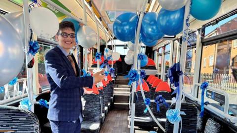 A young man dressed in a smart navy suit and glasses stands in an empty bus with blue and grey striped seats. The bus is decorated with blue and white balloons and ribbons.