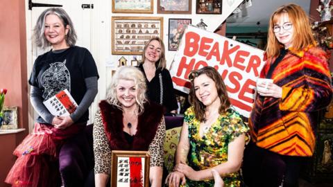 Beaker, a band consisting of five women, pose for a picture in the room of a house. To the right, a redheaded woman with glasses and a colourful orange coat, holds a sign that reads "BEAKER HOUSE" in big red letters. She and the others smile in the direction of the camera. 