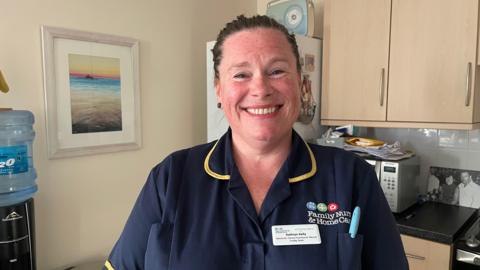 The image shows a woman in a nurse's uniform standing in a kitchen and smiling at the camera.