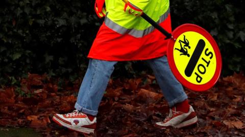 A general view of the feet of a school crossing patrol man, known as a Lollipop Man, carrying a sign that reads: STOP and has the image of two children underneath.