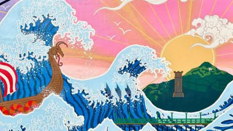 A brightly coloured mural covers the gable end of a building, depicting towering Japanese-inspired waves carrying a Viking longboat past Ramsey landmarks including Queen’s Pier, Albert Tower and North Barrule. A glowing sunrise and stylised clouds create a vibrant backdrop, reimagining Hokusai’s The Great Wave in an unmistakably Manx setting