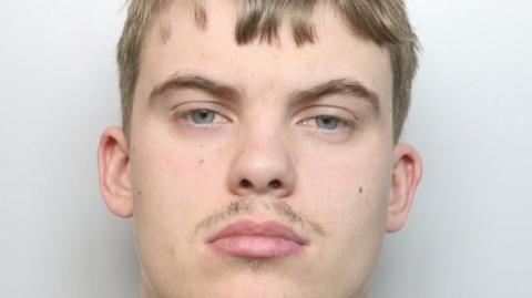 A man with light facial hair and short hair is wearing a grey jumper.
