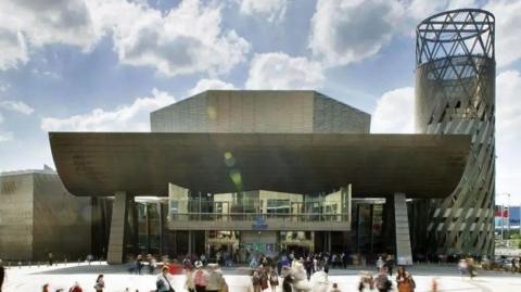 The Lowry arts centre, a steel and glass-clad building facing a paved public plaza. Crowds can be seen milling around the building.