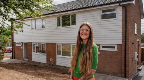 A woman smiling in front of a red brick and white-clad semi-detached house. She is wearing glasses and a green top.
