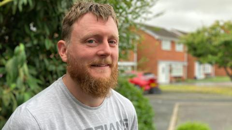 A man with a ginger beard and brown hair, wearing a grey t-shirt, standing in front of a tree with a red car and houses in the background.
