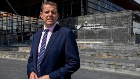 Plaid Cymru's leader Rhun ap Iorwerth standing in front of the Senedd building in Cardiff Bay. He is wearing a navy suit and a pink shirt, with a striped tie.  It is a sunny day.