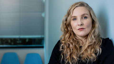 Tamsin McNally looks into the camera. She has long, wavy blonde hair and is wearing a plain black top with blue chairs visible over her right shoulder. 