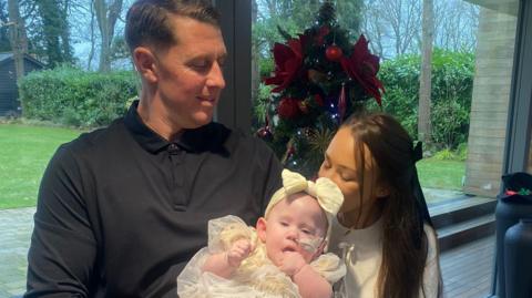 A man and a woman holding a baby girl. He is wearing a black top and she is wearing a white top. The baby is wearing a cream dress and matching bow. A decorated Christmas tree is in the background.