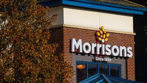 The exterior of a Morrisons on a sunny day, with the logo prominent