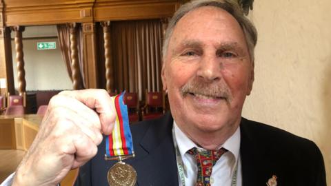 Alan Dowson, who has short grey hair and a moustache, smiling into camera holding up Nuclear Test Medal. He is wearing a navy suit, white shirt and red-patterned tie.