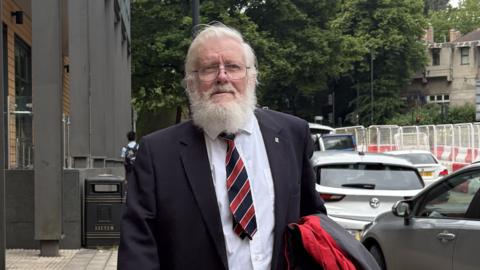 An old man with white grey hair, beard and moustache wearing glasses and a suit with a stripy tie is walking down a road in a city centre, next to a row of cars.