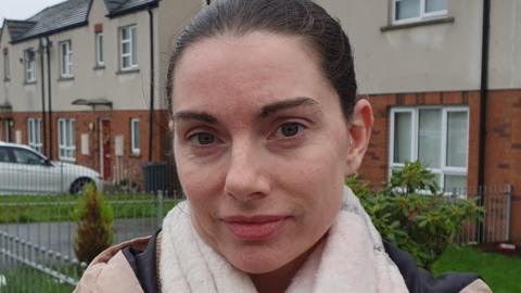 A woman whose hair is dark and tied back stands in front of a row of houses. She is wearing a pink coat and a white wollen scarf