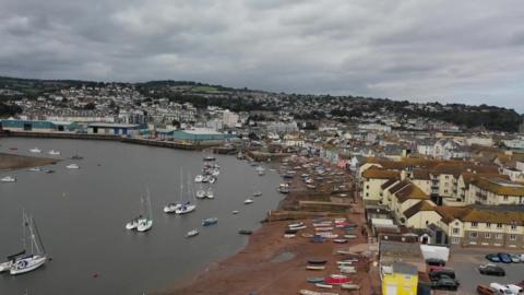 Drone shot showing a beach with boats on, wit more boats in the sea, and houses behind the beach