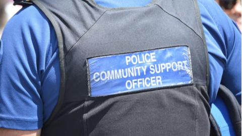 A picture of the back of a officer. It has a sign on the back of the black vest which says police community support officer.